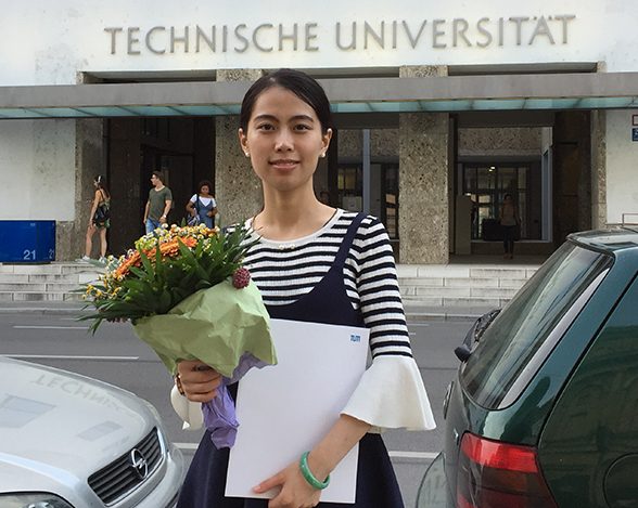 TUM Alumna Yijia Zhuang with her graduation certificate and a bouquet of flowers in from of the TUM main entrance.