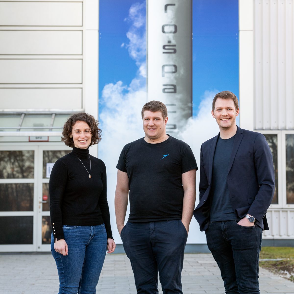 Porträtbild von Lucrezia Veggi, Josef Fleischmann und Daniel Metzler vor den Produktionhallen von Isar Aerospace.