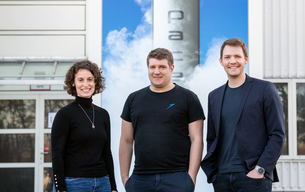 Porträtbild von Lucrezia Veggi, Josef Fleischmann und Daniel Metzler vor den Produktionhallen von Isar Aerospace.