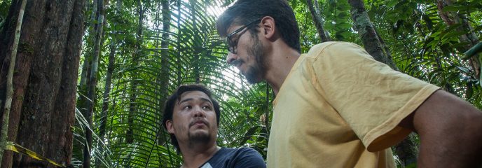 David Lapola in the yellow T-shirt talks to a staff member holding a laptop in the jungle.