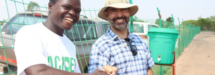 Zwei Männer stoßen freundschaftlich ihre Fäuste gegeneinander, um ihre Zusammenarbeit bei landwirtschaftlichen Projekten in Malawi zu besiegeln. Beide lächeln. Der Hintergrund zeigt eine einfache Wasserstation.
