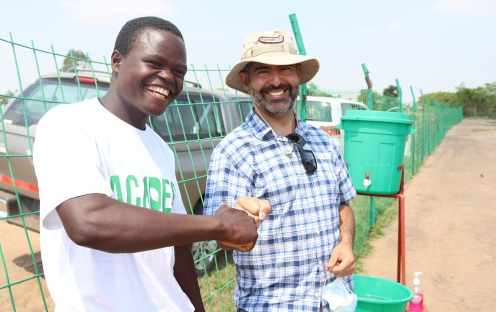 Zwei Männer stoßen freundschaftlich ihre Fäuste gegeneinander, um ihre Zusammenarbeit bei landwirtschaftlichen Projekten in Malawi zu besiegeln. Beide lächeln. Der Hintergrund zeigt eine einfache Wasserstation.