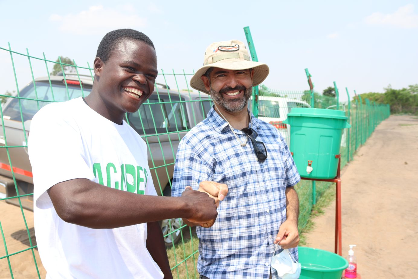 Zwei Männer stoßen freundschaftlich ihre Fäuste gegeneinander, um ihre Zusammenarbeit bei landwirtschaftlichen Projekten in Malawi zu besiegeln. Beide lächeln. Der Hintergrund zeigt eine einfache Wasserstation.